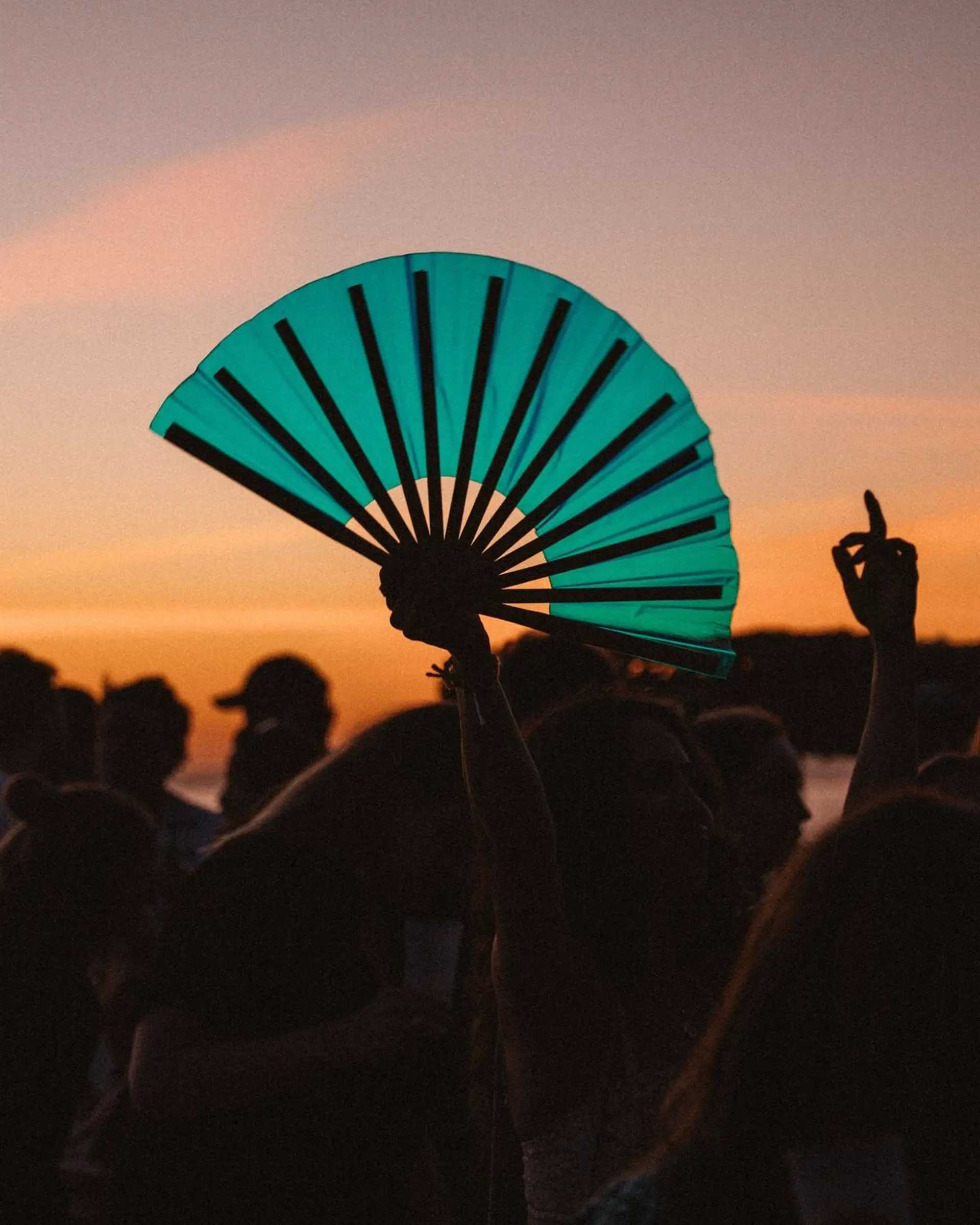 A dancer holds a green hand fan open above a crowd silhouetted against an orange sunset.